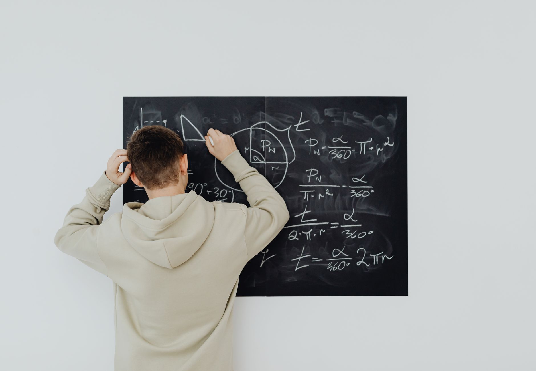 Teenager in hoodie writing math formulas on a blackboard indoors, showcasing problem-solving skills.