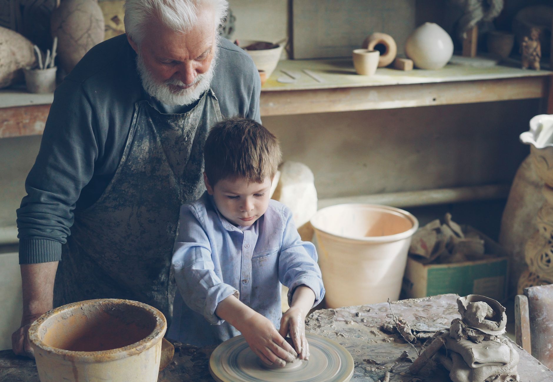 Elderly man guiding young boy in pottery making in rustic workshop.