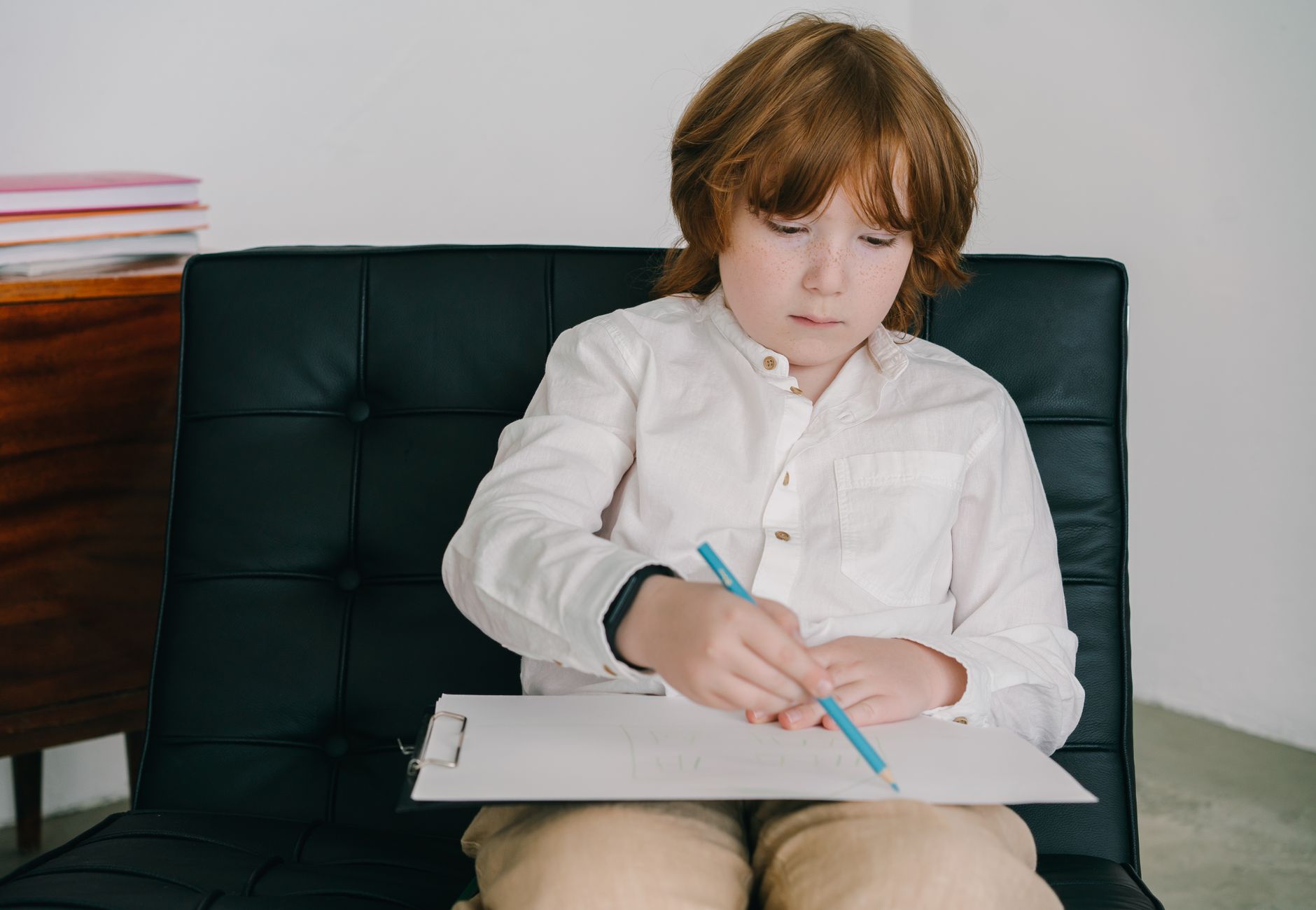 Child with red hair concentrates on drawing with colored pencils while sitting on a black chair.